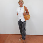 Woman wearing a white sleeveless top and patterned pants, holding a brown bag, standing on a tiled floor.