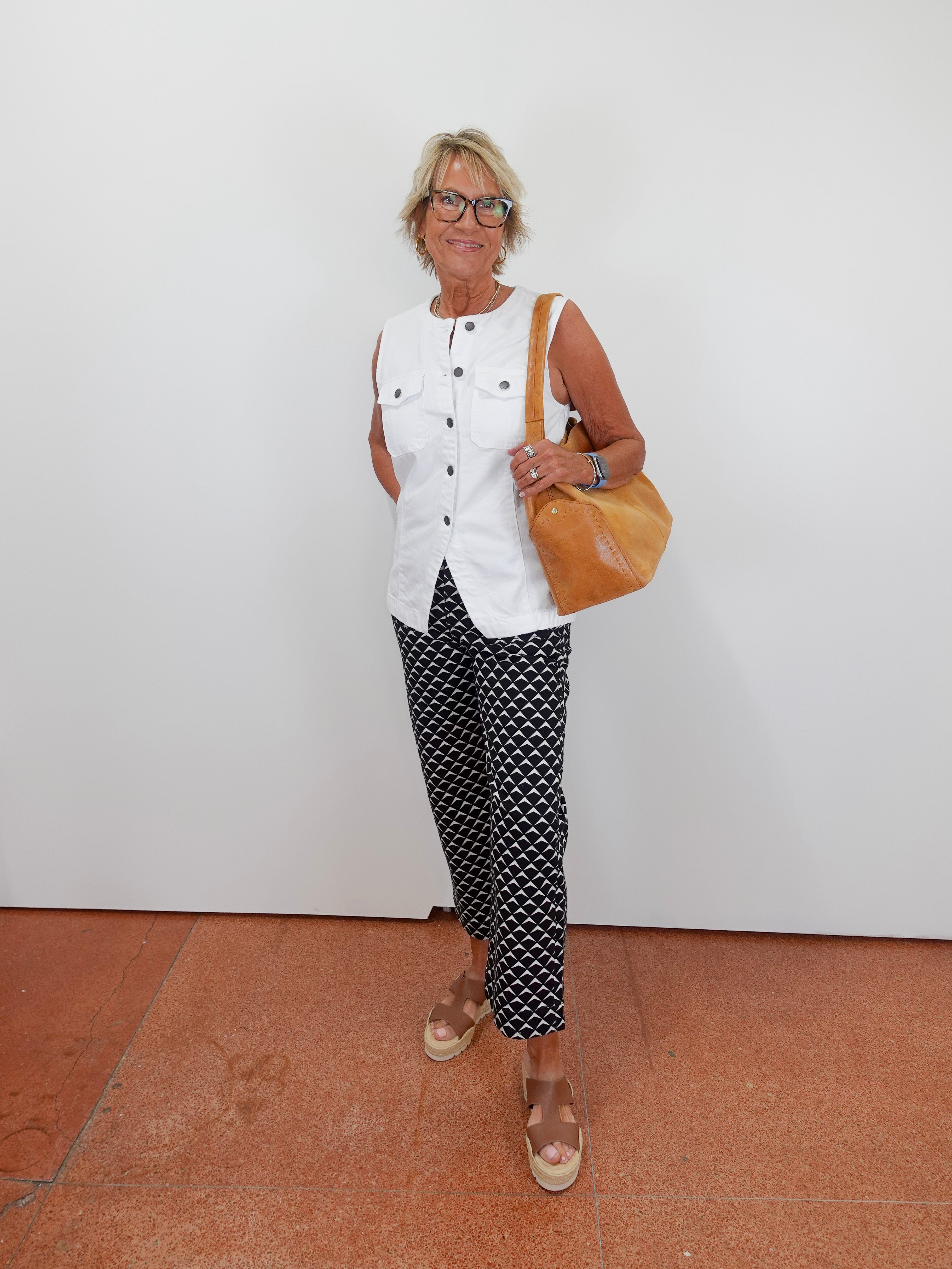 Woman wearing a white sleeveless top and patterned pants, holding a brown bag, standing on a tiled floor.