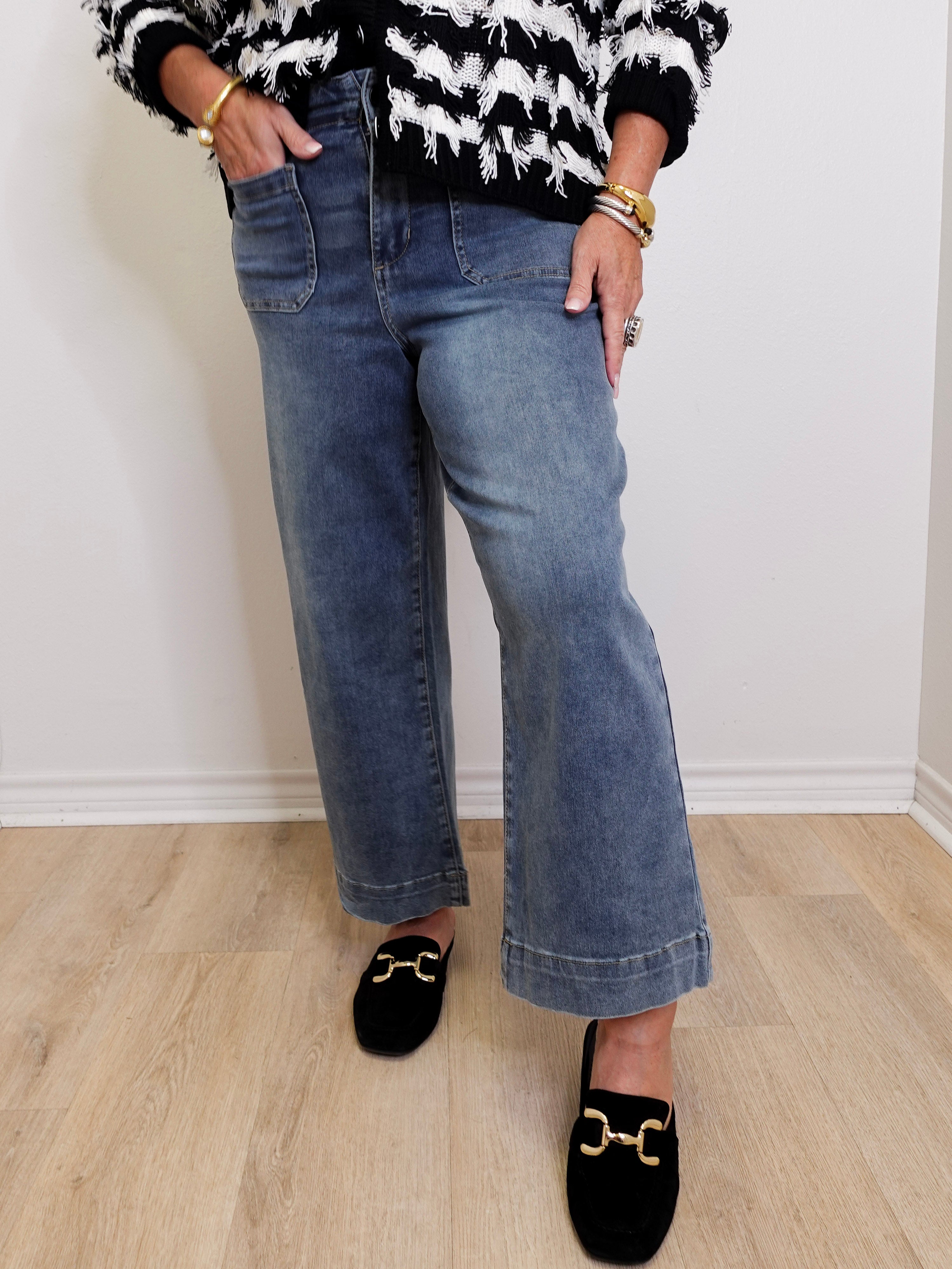 Person wearing a black and white patterned sweater, blue jeans, and black slippers on a light wooden floor.