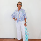 Person wearing a striped shirt and white pants, holding a blue and white striped bag against a white wall.