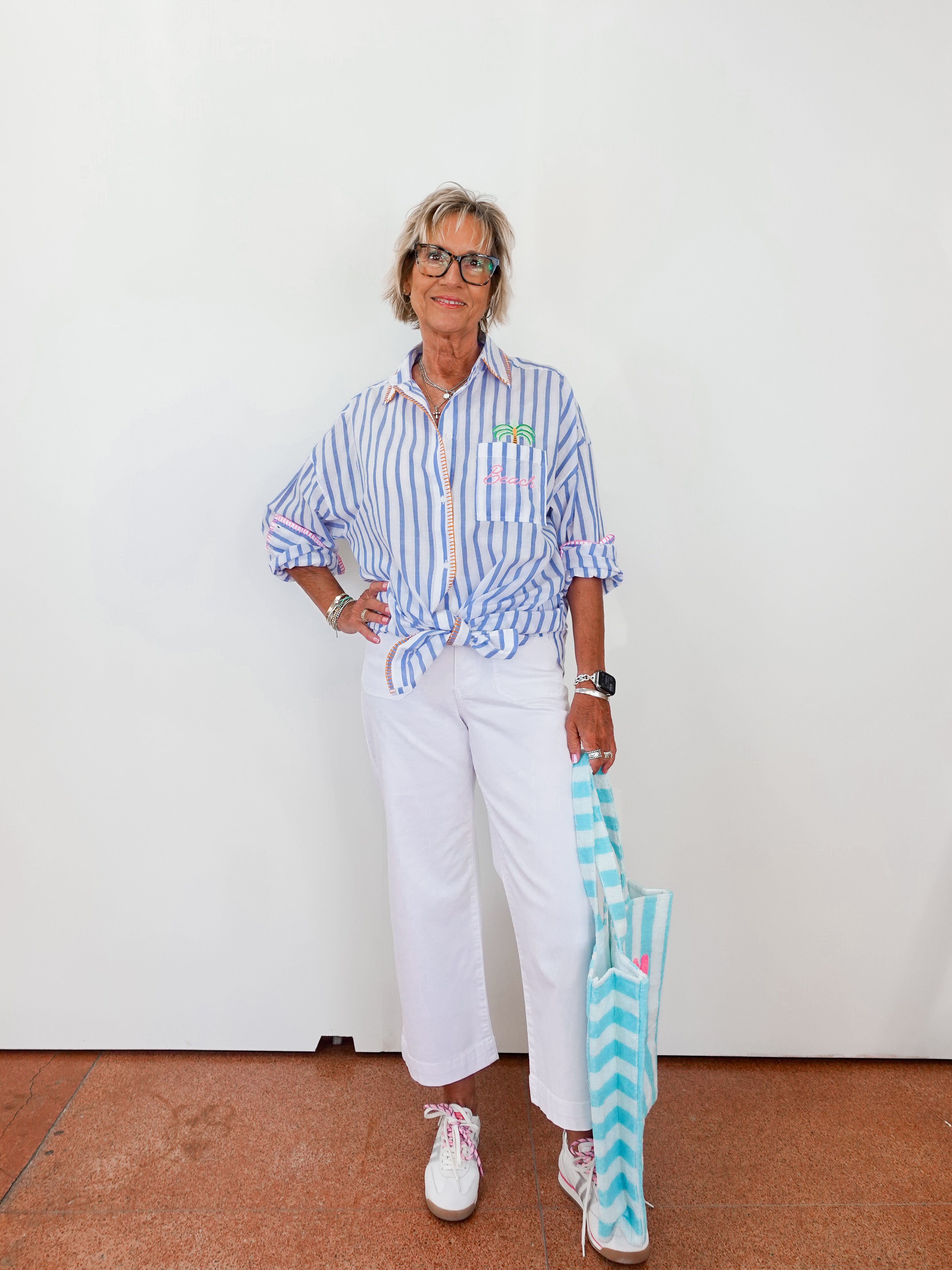 Person wearing a striped shirt and white pants, holding a blue and white striped bag against a white wall.