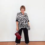 Woman wearing a black and white patterned top with black pants, holding a red handbag against a white background.