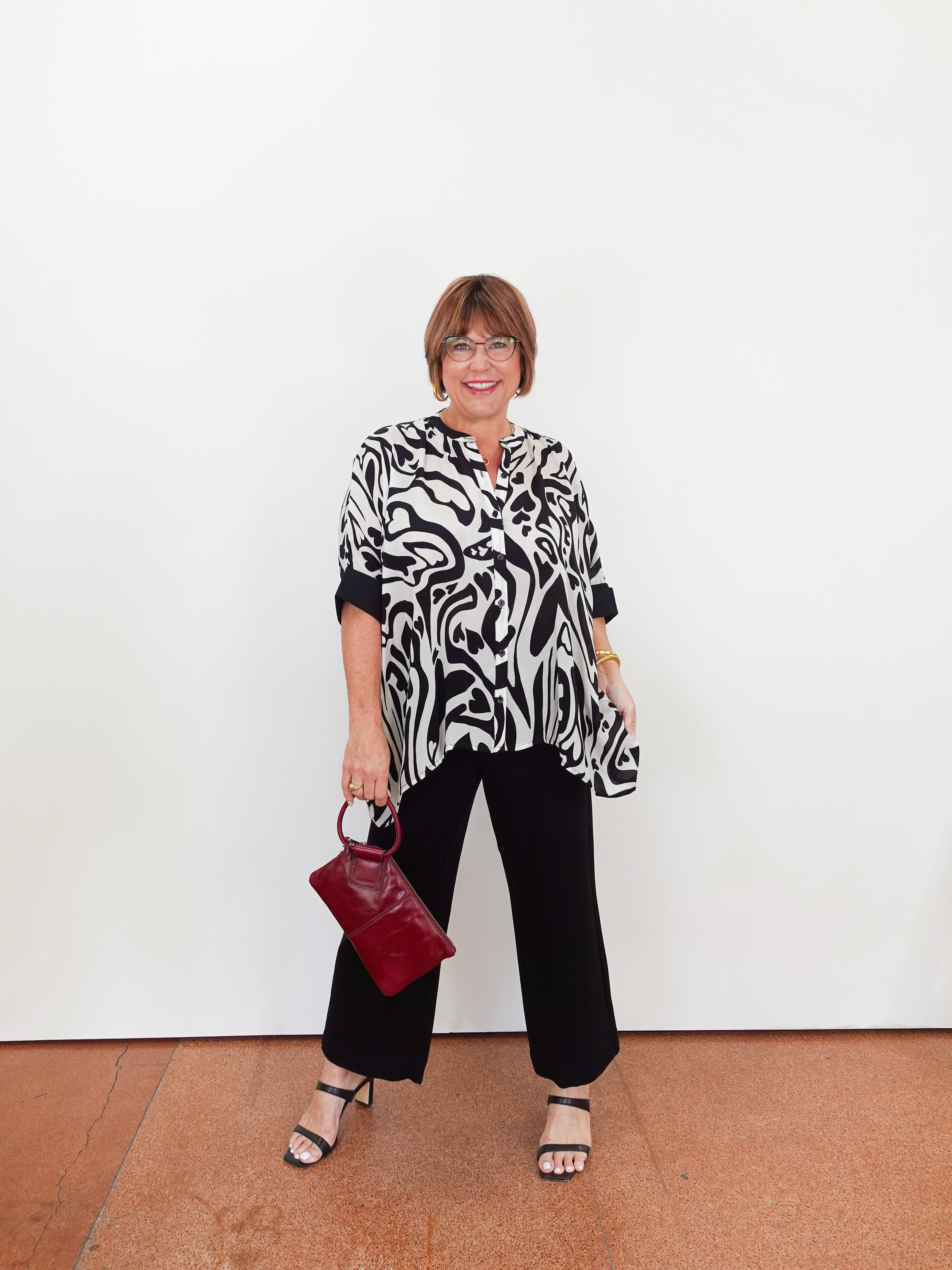 Woman wearing a black and white patterned top with black pants, holding a red handbag against a white background.