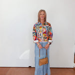 Woman wearing a colorful floral blouse and blue jeans, standing against a white wall.