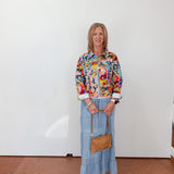Woman wearing a colorful floral blouse and blue jeans, standing against a white wall.