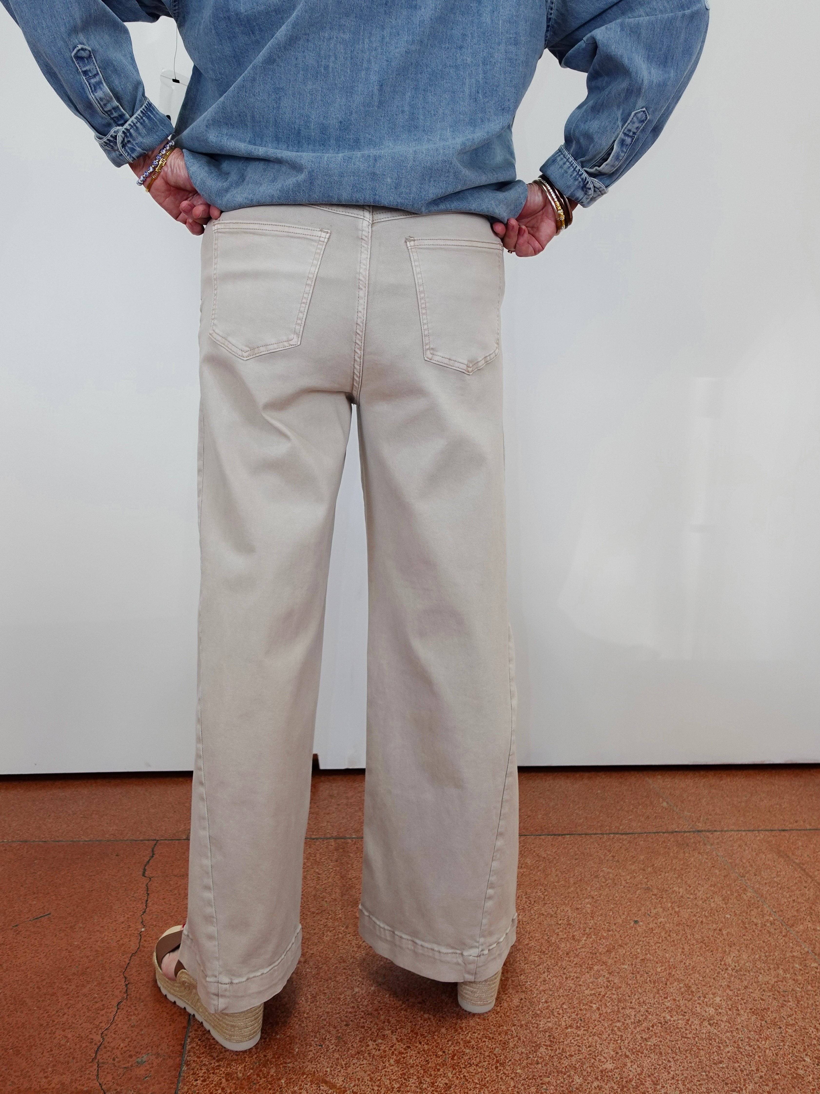 A person shown from the back wearing light beige wide-leg jeans, a blue denim shirt, and beige sandals, standing against a white wall on a terracotta-colored floor.