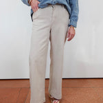 A person wearing light beige wide-leg pants, a blue denim shirt, and patterned platform sandals, standing against a white wall on a terracotta-colored floor.