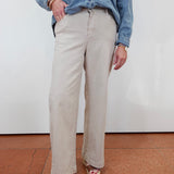 A person wearing light beige wide-leg pants, a blue denim shirt, and patterned platform sandals, standing against a white wall on a terracotta-colored floor.