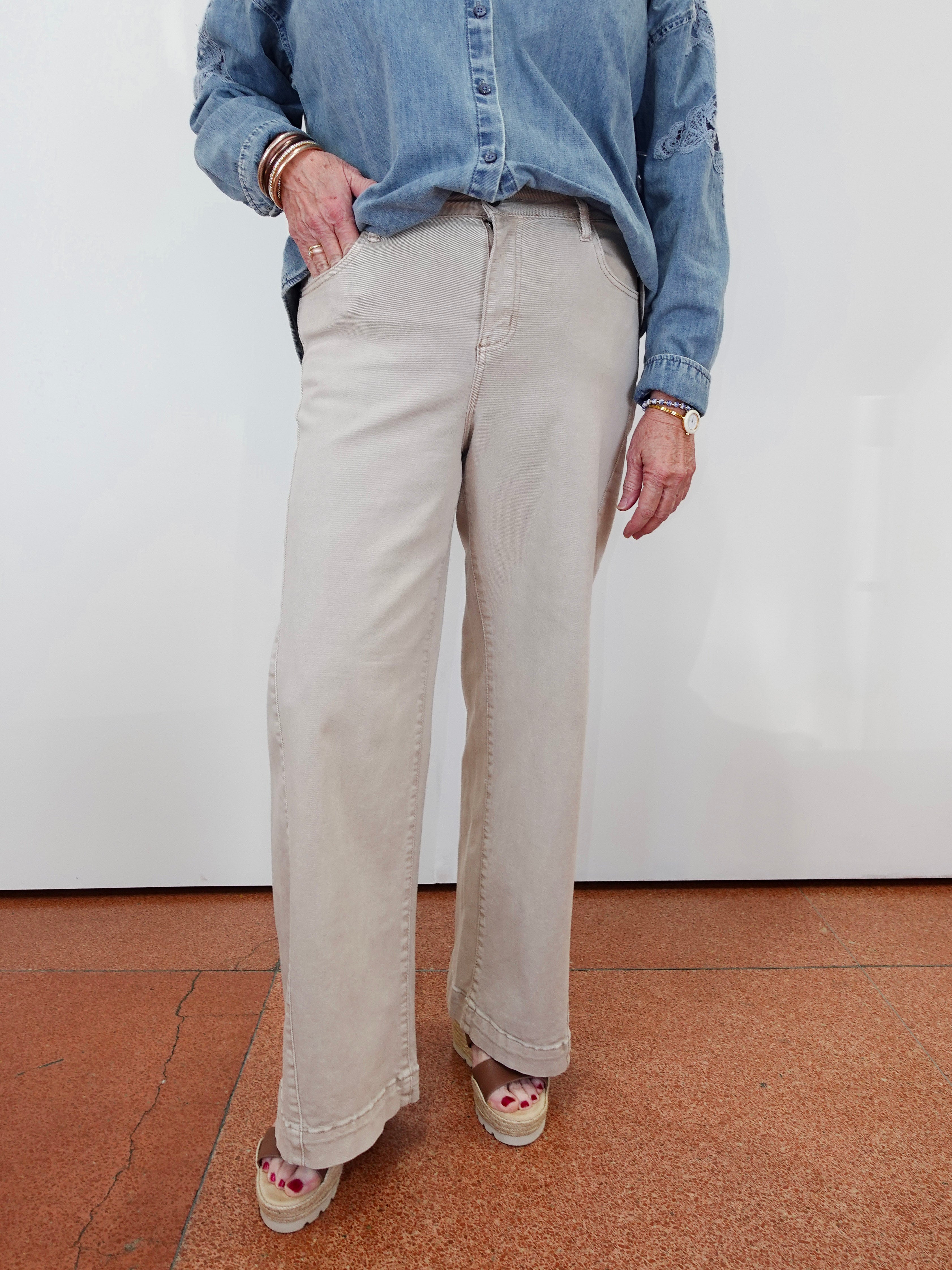 A person wearing light beige wide-leg pants, a blue denim shirt, and patterned platform sandals, standing against a white wall on a terracotta-colored floor.