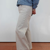 A person shown from the back wearing light beige wide-leg pants, a blue denim shirt, and beige platform sandals, standing against a white wall on a terracotta tiled floor.