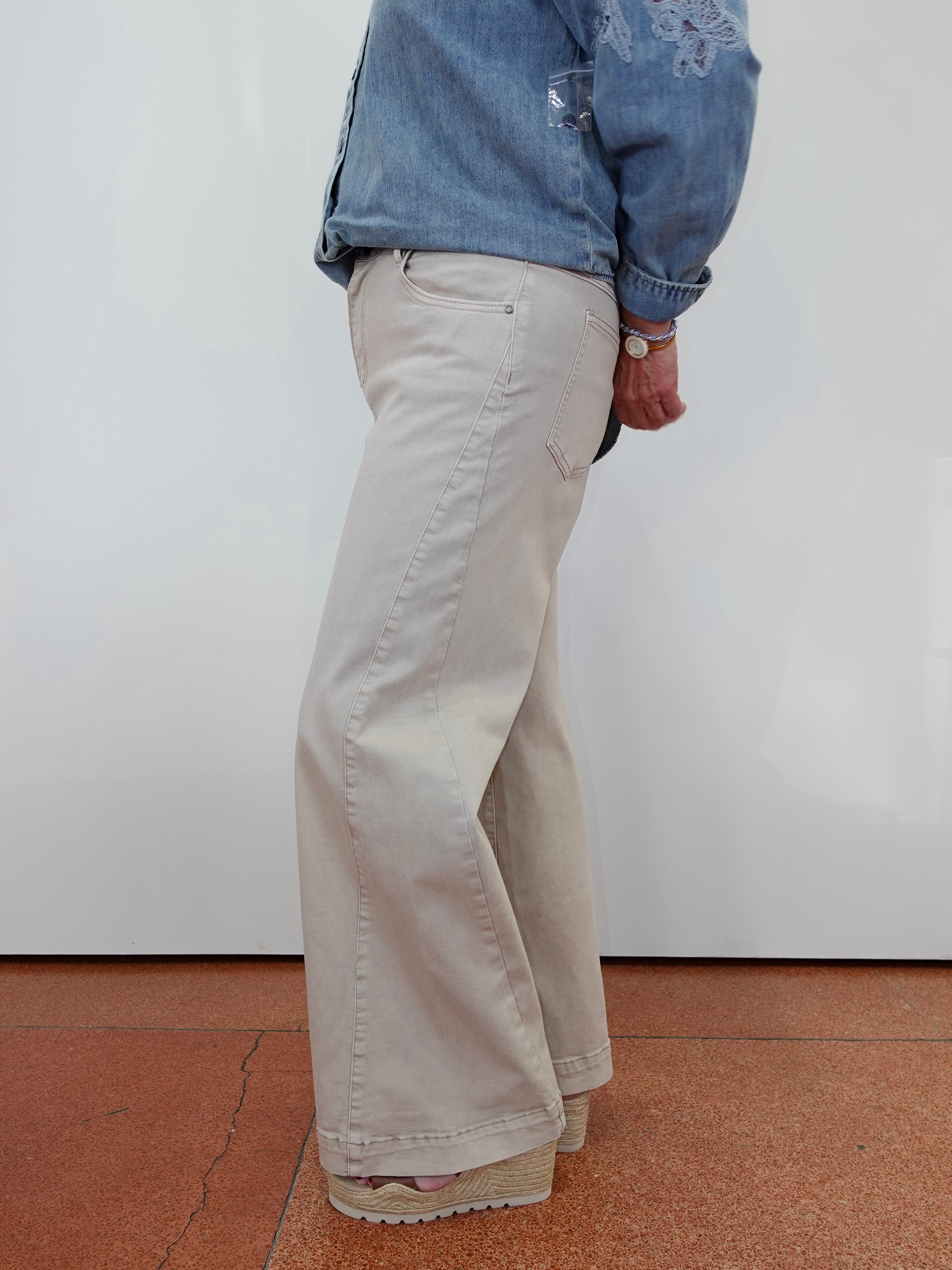 A person shown from the back wearing light beige wide-leg pants, a blue denim shirt, and beige platform sandals, standing against a white wall on a terracotta tiled floor.