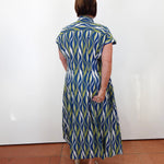 A woman viewed from behind wearing a short-sleeve midi shirt dress with a blue, green, and white wavy pattern, standing against a plain white wall on a terracotta tiled floor.