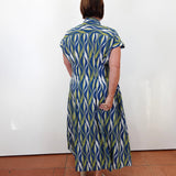 A woman viewed from behind wearing a short-sleeve midi shirt dress with a blue, green, and white wavy pattern, standing against a plain white wall on a terracotta tiled floor.