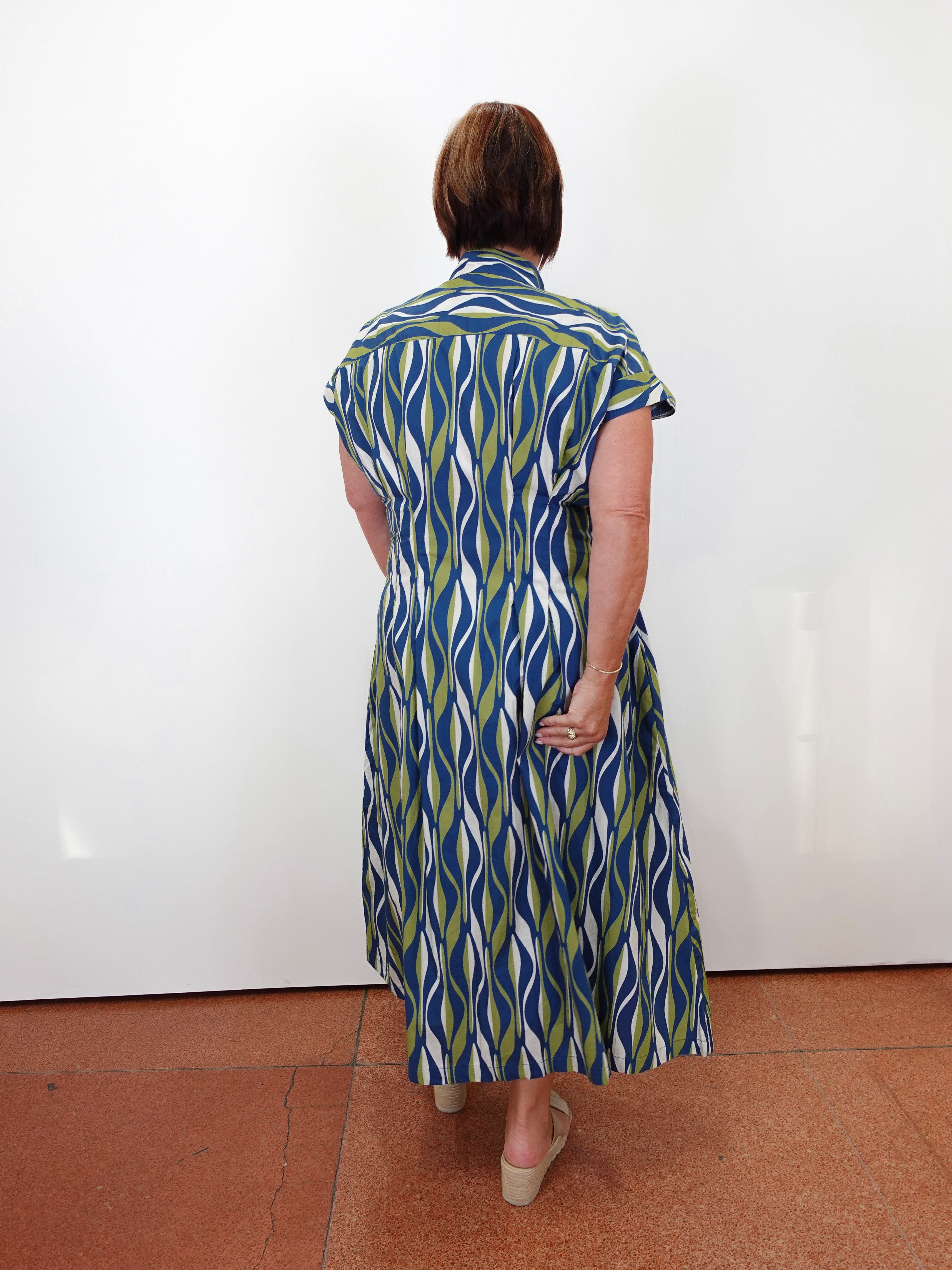 A woman viewed from behind wearing a short-sleeve midi shirt dress with a blue, green, and white wavy pattern, standing against a plain white wall on a terracotta tiled floor.