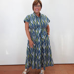 A woman wearing a blue, green, and white patterned midi dress with a tie waist, standing against a plain white wall on a terracotta tiled floor.
