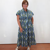 A woman wearing a blue, green, and white patterned midi dress with a tie waist, standing against a plain white wall on a terracotta tiled floor.