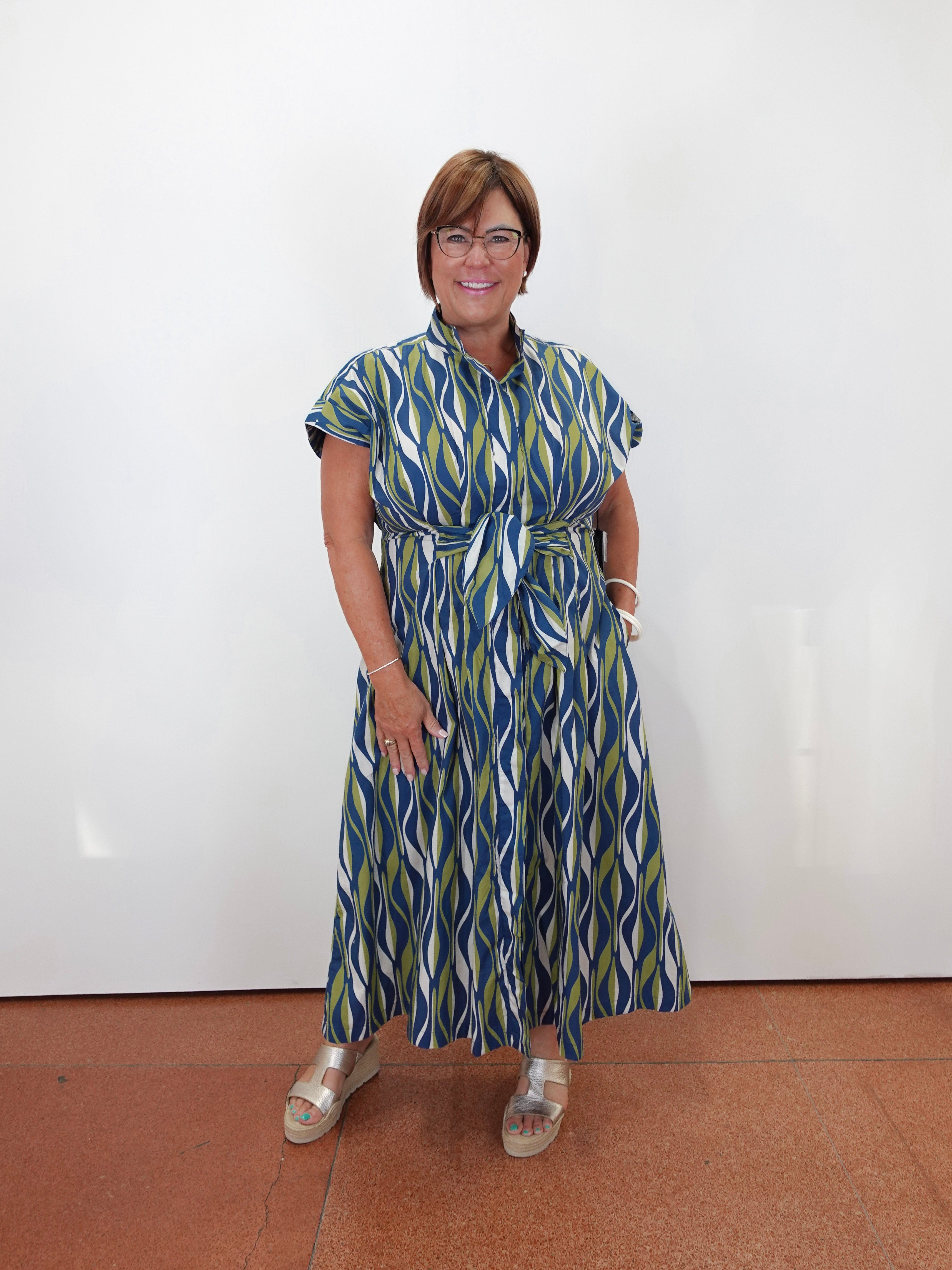 A woman wearing a blue, green, and white patterned midi dress with a tie waist, standing against a plain white wall on a terracotta tiled floor.
