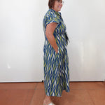 A woman wearing a blue, green, and white patterned midi dress with short sleeves, a tie waist, and side pockets, standing in profile against a white wall on a terracotta tiled floor.