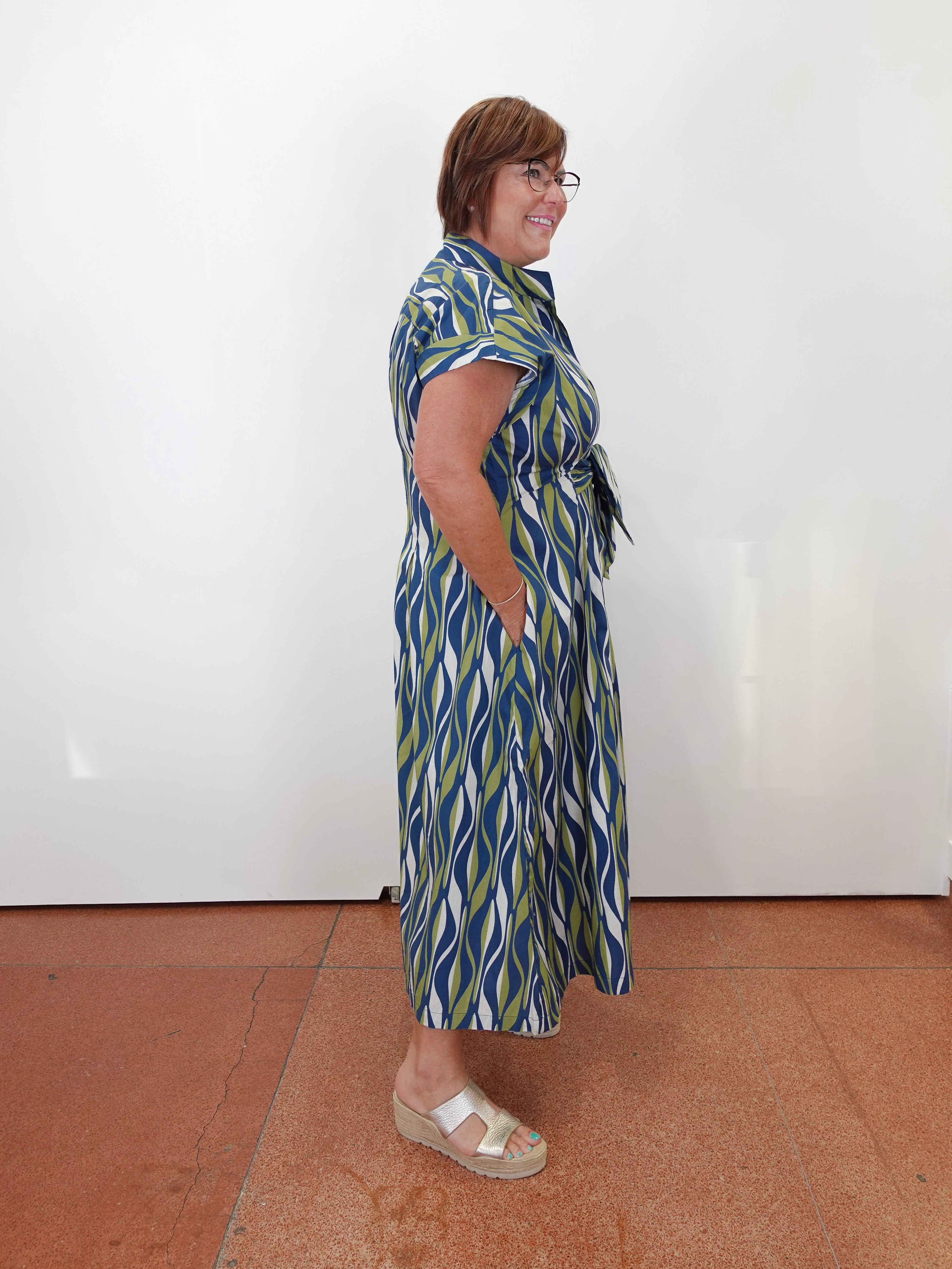 A woman wearing a blue, green, and white patterned midi dress with short sleeves, a tie waist, and side pockets, standing in profile against a white wall on a terracotta tiled floor.