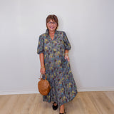 Woman wearing a floral dress holding a brown handbag against a white wall.