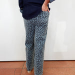 A person wearing blue-gray leopard print pants and a dark navy shirt, standing against a white wall on a terracotta tiled floor.