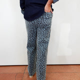A person wearing blue-gray leopard print pants and a dark navy shirt, standing against a white wall on a terracotta tiled floor.