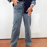A person wearing wide-leg blue-gray leopard print jeans, a dark navy denim shirt, and tan wedge sandals, standing against a white wall.