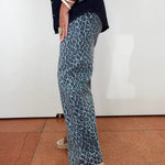 A person wearing wide-leg trousers with a blue and gray leopard print, styled with a dark navy shirt and beige espadrille sandals, standing against a white wall.