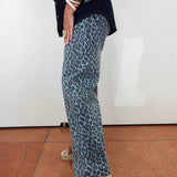 A person wearing wide-leg trousers with a blue and gray leopard print, styled with a dark navy shirt and beige espadrille sandals, standing against a white wall.