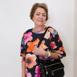 Woman wearing a colorful floral top holding a black handbag against a white background