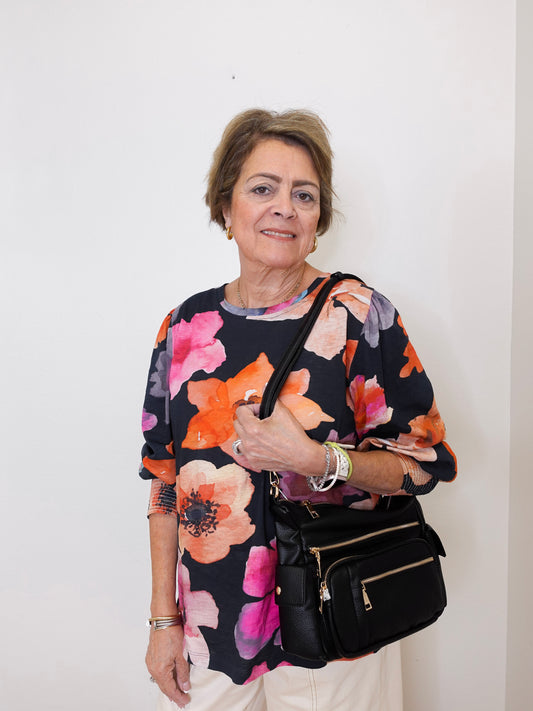 Woman wearing a colorful floral top holding a black handbag against a white background