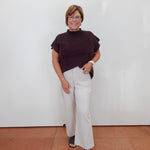 A woman modeling a dark brown short-sleeve mock neck top and off-white wide-leg trousers, standing against a plain white wall with terracotta-colored tiled flooring.