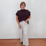 A woman modeling a dark brown short-sleeve mock neck top and off-white wide-leg trousers, standing against a plain white wall with terracotta-colored tiled flooring.