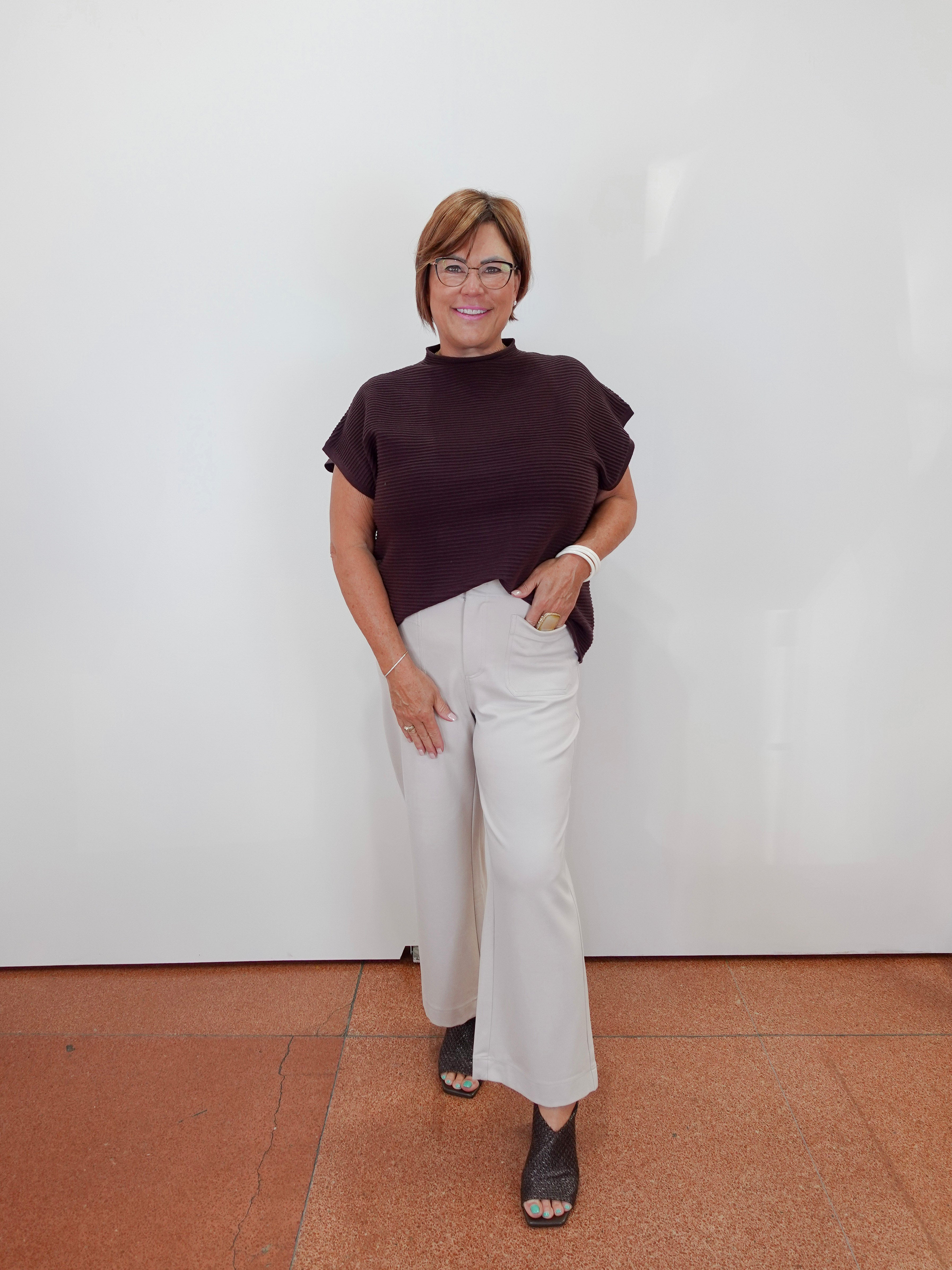 A woman modeling a dark brown short-sleeve mock neck top and off-white wide-leg trousers, standing against a plain white wall with terracotta-colored tiled flooring.