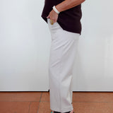A woman shown from the side wearing a dark ribbed sleeveless top and white wide-leg pants, standing against a plain white wall on a terracotta tiled floor.