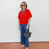 Woman wearing a red top and blue jeans, holding a black handbag against a white wall.