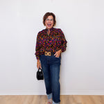 Woman wearing a colorful blouse and blue jeans against a white wall.
