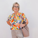 Woman wearing a colorful floral blouse holding a beige handbag against a white background
