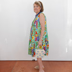 Woman wearing a colorful dress with a pattern of birds and plants, standing against a white wall.