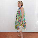 Woman wearing a colorful dress with a pattern of birds and plants, standing against a white wall.