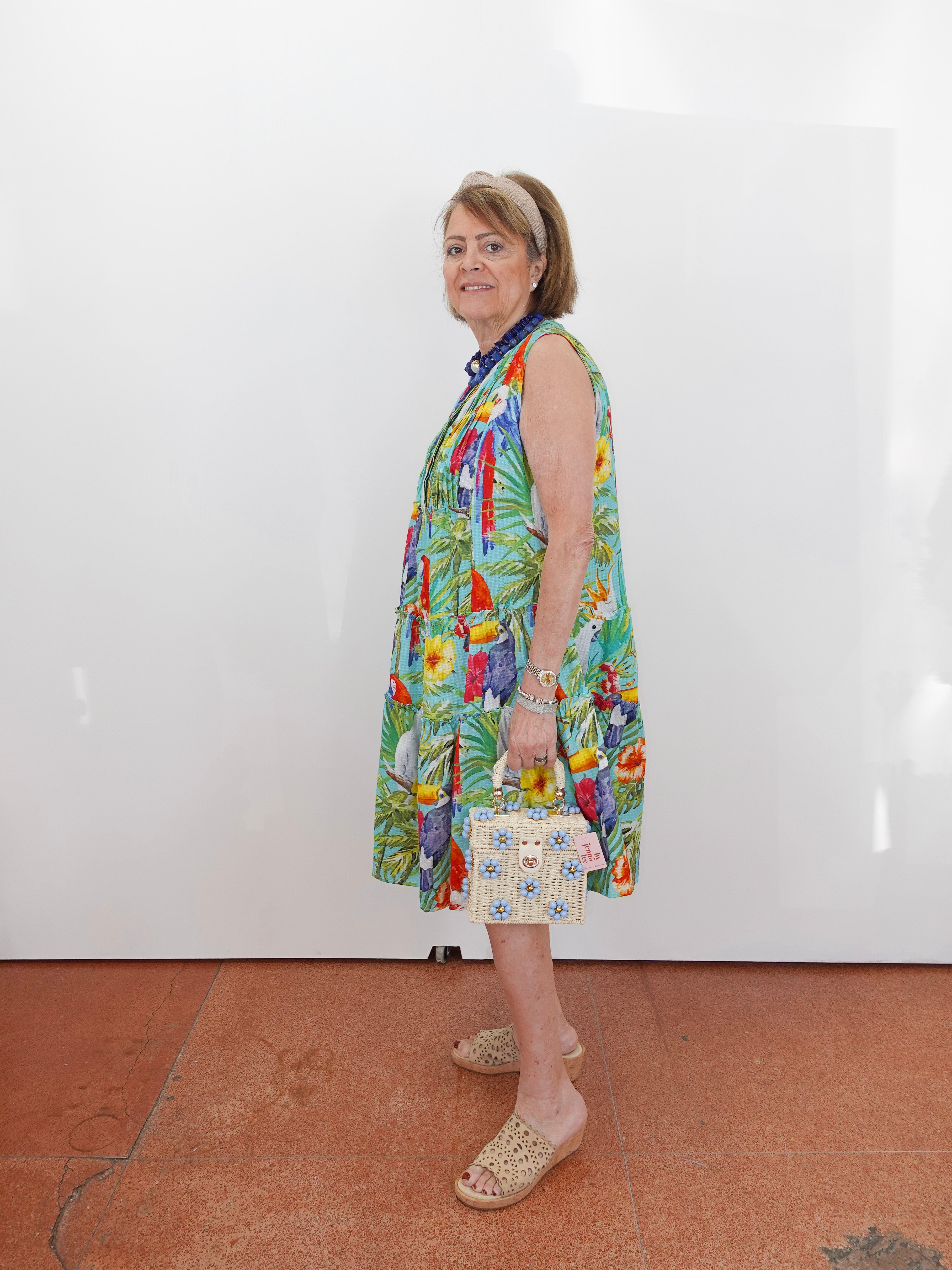 Woman wearing a colorful dress with a pattern of birds and plants, standing against a white wall.