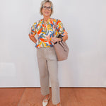Woman wearing a colorful blouse and beige pants, holding a handbag against a white wall.