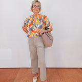 Woman wearing a colorful blouse and beige pants, holding a handbag against a white wall.