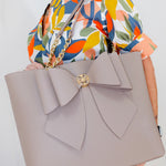 Gray handbag with a large bow held by a person wearing a colorful floral dress.