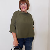 Woman wearing an olive green top and blue jeans, holding a black handbag against a white wall.