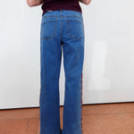 A person viewed from the back wearing blue wide-leg jeans with patterned side panels and a dark ribbed top, standing against a white wall on a terracotta floor.