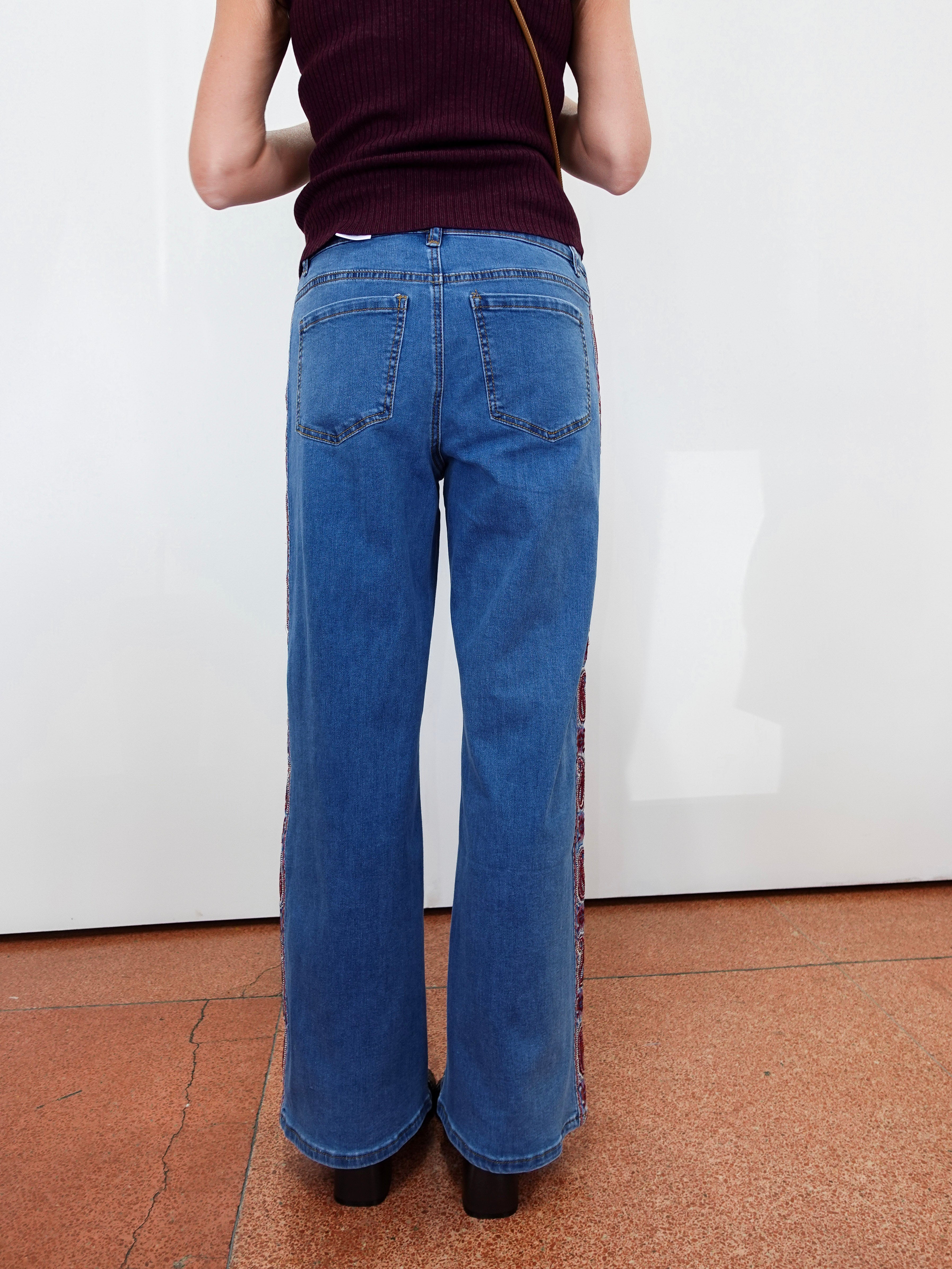 A person viewed from the back wearing blue wide-leg jeans with patterned side panels and a dark ribbed top, standing against a white wall on a terracotta floor.