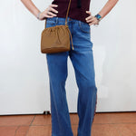 A person wearing blue wide-leg jeans with side cutouts, a dark burgundy ribbed top, and black sandals, carrying a brown leather bucket bag with a gold chain strap against a white wall.
