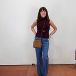 A woman stands against a plain white wall wearing a sleeveless burgundy mock-neck top, blue wide-leg jeans, and black sandals, while carrying a brown crossbody bag.