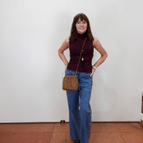 A woman stands against a plain white wall wearing a sleeveless burgundy mock-neck top, blue wide-leg jeans, and black sandals, while carrying a brown crossbody bag.